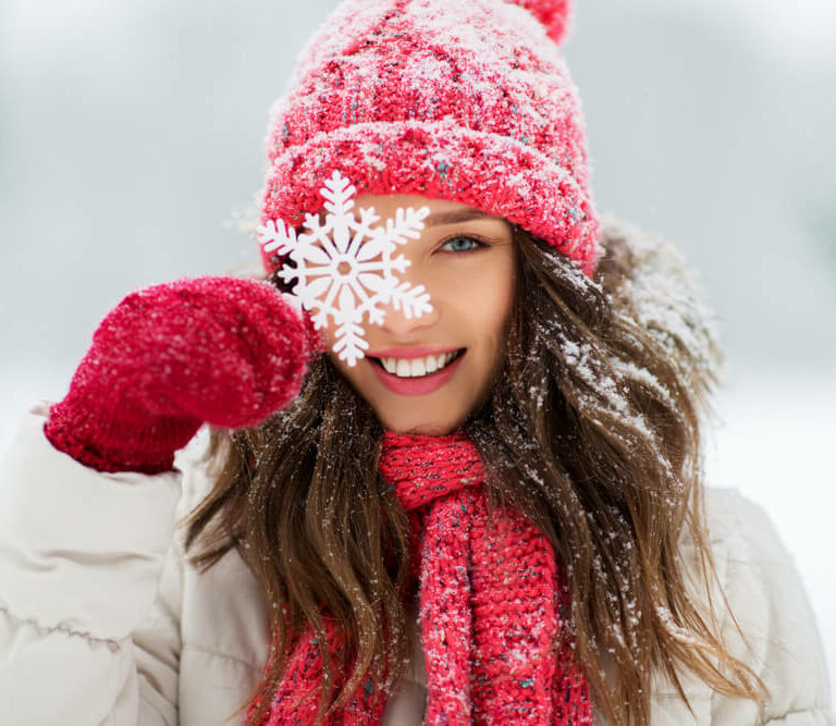 Image shows woman smiling in winter clothes with snow in the background holding giant snowflake in front of her eye.