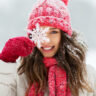Image shows woman smiling in winter clothes with snow in the background holding giant snowflake in front of her eye.