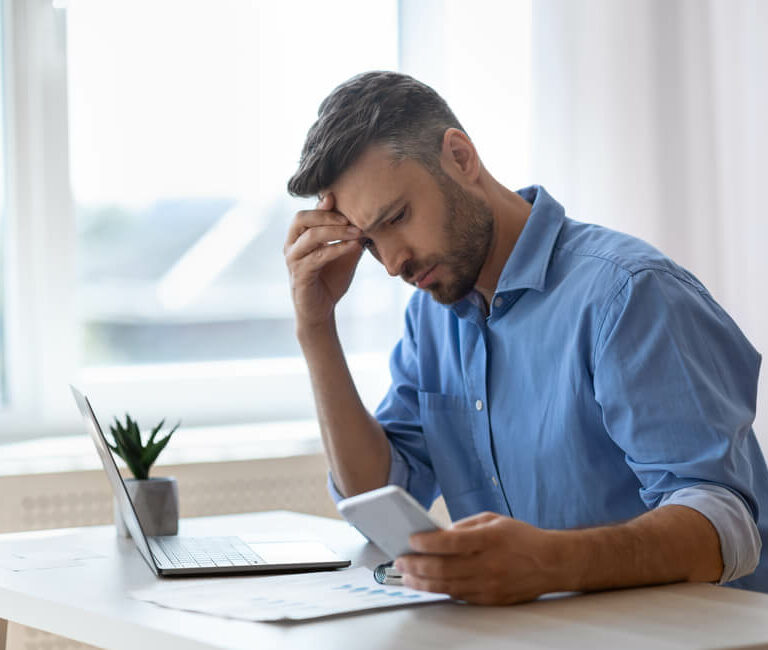 Image shows man sitting at desk looking down at phone with strained eyes