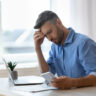 Image shows man sitting at desk looking down at phone with strained eyes